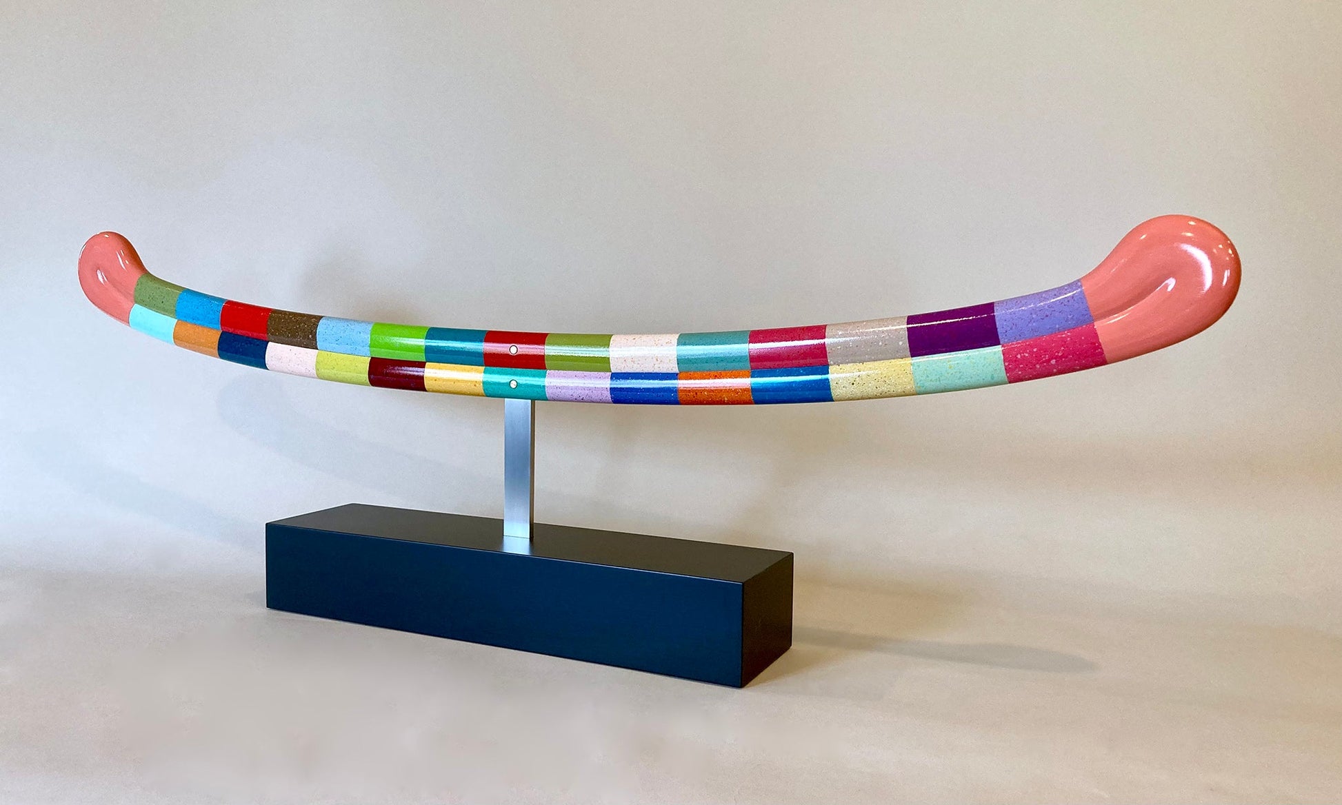 Abstract sculpture depicting a closed mouthed smile. The smile consists of 30 different colors arranged in rectangles along the lips. It is suspended with a silver bar over a black block. 13"x40"x4.25" painted wood by Sean O'Meallie