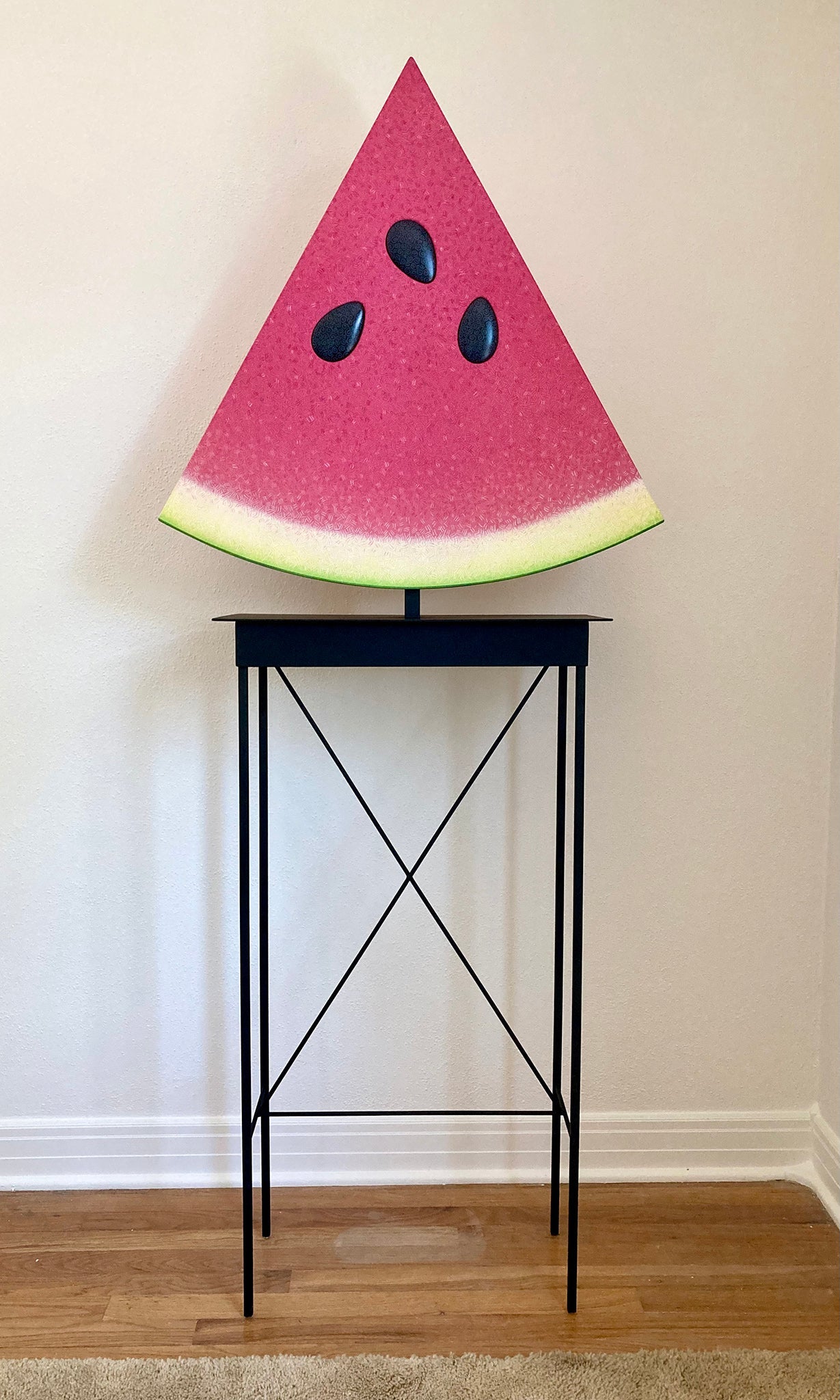 Sculpture depicting a large slice of watermelon atop a black table. 2022 70"x28"x10" painted wood, steel table by Sean O'Meallie