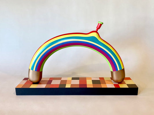 Wooden sculpture depicting a balloon in the shape and painted to look like a rainbow. The ends of the balloon are gold. The balloon sits atop a board that is painted in different shades of red and yellow. 2022 16"x26.75"x5.5" painted wood by Sean O'Meallie